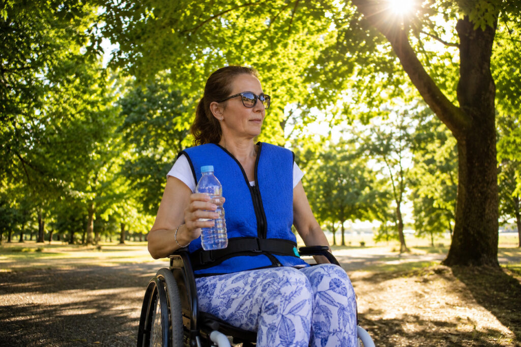 Frau im Rollstuhl mit Kühlkleidung, Kühlweste und Wasserflasche in der Hand.