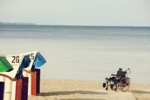Ein Rollstuhl steht am STrand im Sand. Daneben 3 Strandkörbe. Im Hintergrund das Meer.